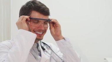 İyi aydınlatılmış bir laboratuvarda, koyu saçlı ve sakallı neşeli bir erkek doktor şeffaf gözlük takıyor, sıcaklık ve profesyonellik saçıyor. Arkadaşça gülümsemesi ve beyaz önlüğü genç hastaları rahatlatmak için mükemmel bir ortam yaratıyor.