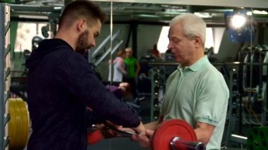 Genç bir erkek antrenör, parlak kırmızı bir halteri kendinden emin bir şekilde aynanın önünde kaldıran üst düzey bir müşteriye hevesle veriyor. Sahne, spor salonunun enerjik enerjisini yakalıyor. Hem antrenörün hem de yaşlı adamın kendini adamışlığını gözler önüne seriyor.