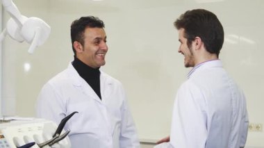 Neşeli olgun bir erkek dişçi, genç meslektaşı ile parlak bir diş kliniğinde sıcak bir şekilde tokalaşıyor, bu da ortaklığı ve sağlık hizmetlerindeki başarıyı simgeliyor. Gülüşleri profesyonellik ve dostluk saçıyor. Gelişen bir tıbbi ekip sergiliyorlar.