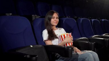 Çekici genç bir kadın rahat bir şekilde lüks bir sinema koltuğunda oturur ve çizgili bir kova patlamış mısırı tutarken yanındaki boş koltuğu işaret eder. Işıl ışıl gülüşü ve rahat tavırları davetkar bir atmosfer yaratıyor. Neşeli bir ortam için mükemmel.