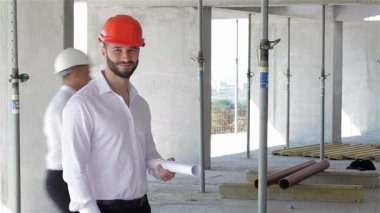 Turuncu bir başlık takan kendine güvenen beyaz erkek müteahhit, hareketli bir inşaat sahasının ortasında mimari planlar yapmaktan gurur duyuyor. Arka planda, iki beyaz adam el sıkışıyor, dinamik dünyada işbirliği ve takım çalışmasını simgeliyor.