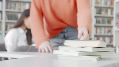 Samimi turuncu süveter giyen odaklanmış bir kız öğrenci kütüphanede ders çalışmak için arkadaşına katılmaya hazır. Sahne, arka planda hafif bir kitap rafı bulanıklığı ile akademik işbirliğinin özünü yakalıyor.