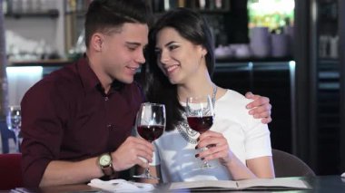 Çekici beyaz bir adam şık bir restoranda esmer kız arkadaşını kırmızı şarapla kadeh kaldırırken kucaklıyor. Neşeli gülümsemeleri ve samimi bakışları sıcak bir atmosfer yaratıyor, modern ortamda aşkı ve bağlantıyı kutluyor.