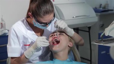 Yetenekli genç bir diş cerrahı, gözlük takan ve maske takan, hastaların dişlerini incelemek için dikkatlice diş ve ayna takıyor. Dişçi koltuğundaki sarışın kadının ağzı sonuna kadar açık, profesyonel ve kısır bir kadın var.