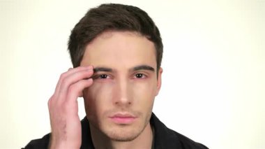 Keskin çene çizgisi olan kendine güvenen genç bir adam aynanın önünde diz çöküp beyaz bir arka plana karşı yakışıklı özelliklerini sergiliyor. Düşünceli bir ifadeyle, nazikçe yüzünü tıraş köpüğüyle silerken, aynı zamanda büyüleyici ve modern stil yayıyor.