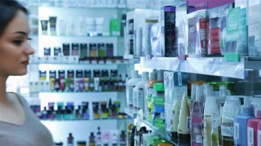 Bir eczanedeki renkli ürünlerin arasında şampuan etiketini okumaya derinden odaklanmış genç bir kadının büyüleyici portresi. Bu sahne güzel bir şekilde sağlık ve güzellik alışverişinin özünü yansıtıyor, düşünceli ifadesini sergiliyor ve