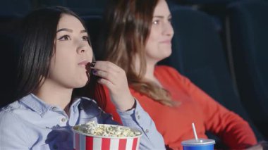 Sıcak bir sinemada, iki genç kadın, patlamış mısır ve gazoz yudumlayarak keyifli bir film deneyimini paylaşıyor. Neşeli ifadeleri filmin heyecanını yansıtıyor, kahkaha ve arkadaşlıkla dolu canlı bir atmosfer yaratıyor.
