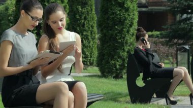 Açık havada şık giyinen iki genç ve çekici iş kadını tabletlerinde anlaşmak için otururken, şık takım elbiseli üçüncü bir kadın da yakınlarda bir telefon alıyor. Bu dinamik sahne modern iş hayatının özünü yansıtıyor.