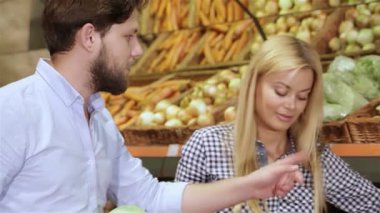 Çekici bir çift canlı bir süpermarketin sebze bölümünde enfes bir alışveriş deneyimi yaşıyor. Sarışın kadın ve kahverengi saçlı adam bir dizi yeni ürünle çevrili renkli bir görüntüyü keşfederken gülümsüyor.