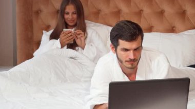 Yakışıklı bir adam dizüstü bilgisayarına odaklanırken güzel karısı arka planda dinleniyor, akıllı telefonuna dalmış. Bu samimi sahne, lüks bornozlar içindeki hoş bir çifti yakalıyor. Lüks otellerinde konforu ve bağlantıyı somutlaştırıyorlar.