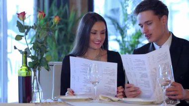 Zarif giyimli genç bir çift, hoş bir restoranın menüsünü incelerken gülümsüyor, yumuşak ışıklandırma ve güzel bir çiçek süsü ile çevrili. Neşeli ifadeleri, kutlamak için mükemmel yemeği seçmenin heyecanını yansıtıyor.