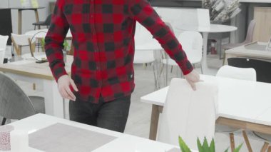 Şık sakallı bir adam şık bir mobilya mağazasındaki çeşitli mobilyaları değerlendirmek için şık bir yemek masasında düşünceli bir şekilde oturur. Çağdaş sandalye ve masalarla çevrelenmiş, modern yaşamın özünü somutlaştırıyor, seçimler yapıyor.
