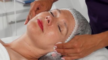 Yetenekli bir kozmetikçi yüzünü yumuşak pamuk pedlerle nazikçe temizleyip cildini gençleştirici bir güzellik tedavisine hazırlarken neşeli bir son sınıf kadını dinleniyor. Huzurlu atmosfer, bu şımartmada gösterilen özen ve profesyonelliği vurguluyor.