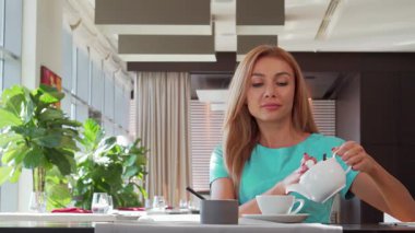 Işıl ışıl saçlı bir kadın fincanına buharlı çay dökerek şık bir kafe ortamının tadını çıkarıyor. Sabah ışığı pencerelerden süzülüyor. Sıcak atmosferi arttırıyor. Yemeğin tadını çıkarıyor, mutluluk ve sükuneti somutlaştırıyor.
