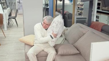 Neşeli yaşlı bir adam şık bir mobilya mağazasında şık bir kanepede otururken şen şakrak bir şekilde yumuşak bir yastığa sarılıyor. Bu canlı sahne, rahat ve boş zamanların özünü yakalar, rahat ev eşyalarını keşfe çıkan erkekleri eğlendirir.