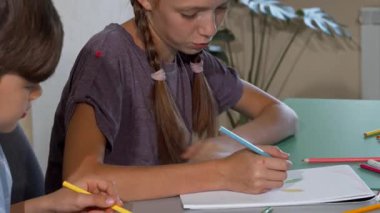 Saçları örgülü odaklanmış genç bir kız ve bir oğlan, renkli kalemler ve parlak, davetkar bir sınıfla çevrili, dersler sırasında resim projeleriyle derinden meşguller. Bu hoş sahne çocukluk yaratıcılığının özünü yansıtıyor.