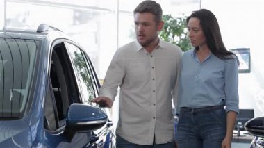 Genç bir çift, galerideki parlak mavi SUV 'yi inceliyor. Beyaz adam, şık sakallı, esmer kız arkadaşı öne eğilip, ortak heyecan ve bağlantılarını gösterirken,