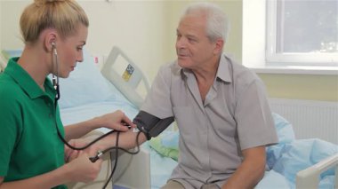 Kendini adamış beyaz bir hemşire, yaşlı bir adamın tansiyonunu dikkatle ölçüyor. Bir hastane yatağında rahatça oturuyor. Hemşire hastaların nabzını dinlemek için steteskop kullanırken, sıcak atmosfer yumuşak ışıklandırmayla gelişir.