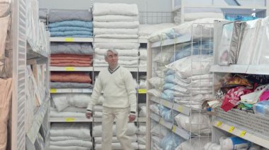 Gümüş saçlı yaşlı bir adam kalabalık bir ev eşyaları mağazasında çeşitli yatak seçeneklerini dikkatle inceliyor. Etrafı yumuşak renklerde tekstil yığınlarıyla çevrili, alışverişin neşesini somutlaştırıyor, rahat dairesini rahatlatmak için.