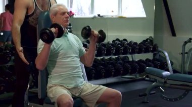 Kendini işine adamış beyaz bir antrenör, dirseklerini tutarak müşterisine nazikçe rehberlik eder, dambılları kaldırırken formda kalmasını sağlar. Yaşlı gri adam, iyi donanımlı bir spor salonunda bir fitness sandalyesine oturmuş, gücünü toplamaya çalışırken azmini sergiliyor.