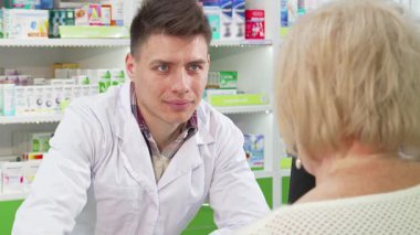 Beyaz laboratuvar önlüğü giymiş genç ve dost canlısı bir kimyager, yaşlı müşterisiyle parlak ve iyi stoklanmış bir eczanede dikkatle meşgul oluyor. Sıcak atmosfer, doğru kişiyi seçmesine yardım ederken şefkatli bir etkileşimin altını çiziyor.