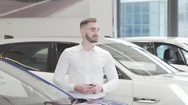 Yakışıklı, mutlu bir adam modern bir araba galerisinde elinde araba anahtarı ve etrafa neşe saçarak gururla durur. Etrafı gösterişli araçlarla çevrili yeni bir otomobil satın almanın heyecanıyla dolu. Başarı ve güven dolu bir yaşam tarzı sergiliyor.