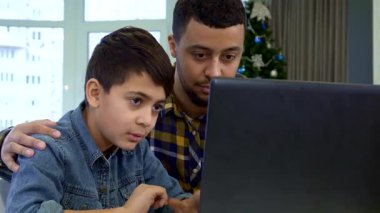 Çekici bir baba ve genç oğlu keyifli bir görüntülü sohbet için dizüstü bilgisayara el sallıyorlar. Sahne, güzel dekore edilmiş bir Noel ağacının karşısında çekiliyor. Ilık ve şenlikli bir neşe saçıyor. Küçük bir çocuk gibi.