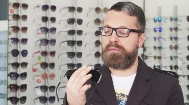 Sakallı bir adam bir optometrist dükkanında durmuş, iki modaya uygun güneş gözlüğü arasında seçeneklerini dikkatlice değerlendiriyor. Renkli gözlüklerle çevrelenmiş, moda ve ultraviyole ışığa karşı keskin bir bakış açısı sergileyen modern tüketiciliğin özünü somutlaştırıyor.