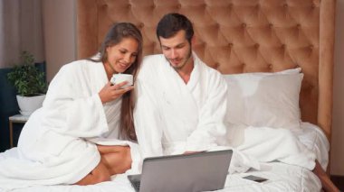 Pofuduk beyaz bornozlu neşeli bir çift yatakta rahat bir anı paylaşıyor, kahvelerini yudumluyor ve dizüstü bilgisayarlarında dijital dünyayı keşfediyorlar. Kahkahaları lüks otel süitini dolduruyor, samimi bir sevgi ve bağlantı ortamı yaratıyor.