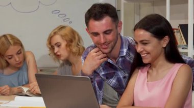 Neşeli genç bir kadın ve erkek sınıf arkadaşı laptopta çalışırken kameraya doğru ışık saçıyorlar. Etrafı odaklanmış arkadaşlarla çevrili. Bu canlı sahne takım çalışmasının ve akademik yoldaşlığın özünü yansıtıyor, öğrenmenin ve öğrenmenin heyecanını gözler önüne seriyor.