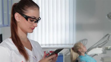 Akıllı telefonunda zeki, modern bir dişçi dolabına güvenle gülümseyen beyaz dişçi tipi genç, çekici bir dişçi. Koruyucu gözlükler ve beyaz bir üniformayla, kadın profesyonellik sergiliyor, hasta ise diş hekiminde dinleniyor.