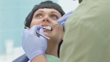 Profesyonel üniforma giymiş yetenekli bir dişçi genç bir kadının dişlerini dikkatle inceliyor. Uzmanlık ve şefkat karışımı bir şey sergiliyor. Parlak, steril klinik ortamı diş sağlığı üzerinde yoğunlaşırken, hastalar