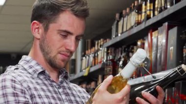 Bakımlı sakallı yakışıklı genç bir adam canlı bir süpermarket koridorunda iki şarap şişesini tutarken kafası karışmış görünüyor. Rengarenk etiketlerle çevrelenmiş, modern tüketici ikilemini somutlaştırıyor.
