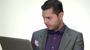 Sivri giyinmiş, papyon takan ve özel dikim takım elbiseli profesyonel bir erkek, dizüstü bilgisayarıyla yakından ilgileniyor, kararlılık ve odaklanma gösteriyor. Temiz beyaz bir arkaplan karşısında verdiği ciddi ifade çalışmalarının yoğunluğunu vurguluyor.