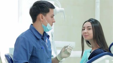 Mavi üniforma ve maske giyen neşeli bir dişçi dişçi, dişçi koltuğunda gülümseyen bir kadın hastayla meşgul olurken hassas aletler seçer. Parlak, steril klinik ortamı profesyonellik ve bakım yayarak rahat bir deneyim sağlıyor.