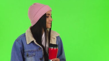 Şık bir kış ceketi ve yumuşak pembe bir şapkayla sarılı hoş bir genç kadın, canlı yeşil bir kromakey arka planına karşı sıcak çayının rahatlatıcı aromasını seviyor. Bu rahat sahne sıcaklık ve neşe saçıyor. Özünü yakalamak için mükemmel.