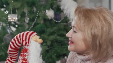 Neşeli, son sınıf öğrencisi bir kadın elinde Noel Baba oyuncağıyla kameraya ışıldıyor. Sıcak ve bayram neşesi saçıyor. Güzel dekore edilmiş bir Noel ağacıyla çevrelenmiş bu şenlik sırasında kutlama ruhunu ve aile sevgisini somutlaştırıyor.