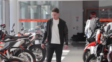 Esmer, beyaz saçlı genç bir adam, bir dizi parlak bisikletle çevrili modern bir motosiklet galerisinde tefekkür ediyor. Nazikçe çenesini eline dayayıp motorsikletlerin çarpıcı tasarımlarını ve canlı renklerini değerlendiriyor.