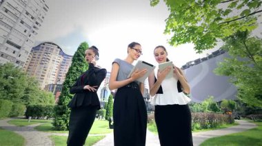 Canlı bir şehir parkında, üç kendine güvenen iş kadını tabletleri üzerinde işbirliği yapıyor, görüşlerini paylaşıyor ve son haberleri tartışıyorlar. Şık resmi kıyafetleri ve ışıl ışıl gülümsemeleri iş dünyasındaki başarılarını ve dostluklarını yansıtıyor.