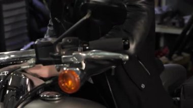 Karizmatik bir erkek bisikletçi, şık bir siyah deri ceketi ve bakımlı bir sakalı olan, hareketli bir atölyede motorsikletinin ön panelini dikkatlice temizlerken gülümsüyor. Yakın plan, parlak krom detaylarını ve tutkusunu yakalar.