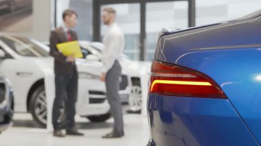 Bir araba satıcısı ve bir erkek müşteri, modern bir otomobil bayiliğinde güven ve ortaklığı simgeleyen sıkı bir tokalaşmaya girişirler. Sahne, ön plandaki mavi bir arabanın parlak çizgileriyle vurgulanarak, yeni sahipliğin heyecanını vurguluyor.