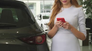 Uçan kızıl saçlı neşeli bir kadın, modern bir araba galerisinde şık bir otomobilin yanında akıllı telefonuyla konuşurken gülümsüyor. Bu canlı sahne araba alışverişinin, teknoloji ve tüketiciliğin heyecanını yansıtıyor.