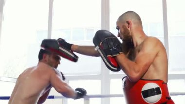 İki kaslı erkek MMA dövüşçüsü, güçlerini ve çevikliklerini sergileyerek dinamik bir antrenman yapıyorlar. Bir dövüşçü güçlü yumruklar atarken diğeri karşılık vermeye hazırlanırken atmosfer enerjiyle doludur.