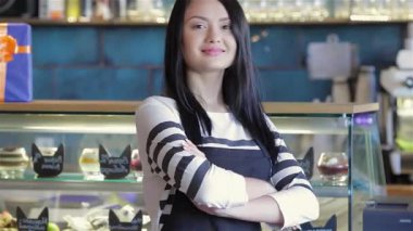 Kendine güvenen genç bir bayan garson kollarını kavuşturup hareketli bir kafede sıcaklık ve cazibe yayıyor. Parlak bir gülümseme ve şık bir önlükle, mükemmel bir servisin özünü temsil ediyor, müşterileri keyifli bir kahveye davet ediyor.