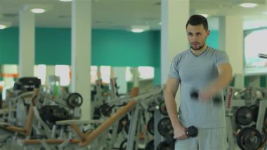 Gri tişörtlü kararlı genç bir adam triseps egzersizlerini dambıllarla yapıyor, gücünü ve bağlılığını parlak bir spor salonunda sergiliyor. Antrenmanından sonra, güvenle koçuna yaklaşır, rehberlik ve geliştirme için hazırdır.