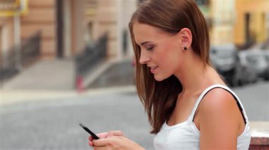 Uzun saçlı şık bir kadın bir bankta oturmuş, canlı bir şehir ortamında akıllı telefonuyla mesajlaşıyor. Güneş sıcacık bir parıltı saçıyor, günlük kıyafetlerini ve etrafındaki hayat dolu sokak manzarasını vurguluyor.