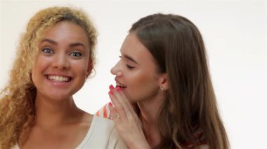 İki genç kadın neşeli bir anı paylaşıyor, bir fısıldayan sırla diğerlerinin kulağına, yumuşak beyaz bir arka plana neşe ve yakınlık yayıyorlar. Kahverengi saçlı Kafkasyalı kız, kızıl saçlı arkadaşı eğildiğinde heyecanla ışıldıyor.