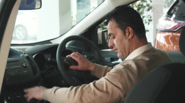 Arka koltuklardan yakalanan koyu saçlı bir adam arabaların özellikleriyle meşgul oluyor, düğmelere basıyor ve dikiz aynasını ayarlıyor. Kameraya sıcak bir gülümsemeyle döner, mükemmel aracı seçmenin heyecanını somutlaştırır.