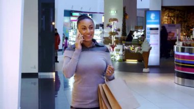 Hareketli bir alışveriş merkezinin hareketli atmosferiyle çevrili bir şekilde cep telefonuyla sohbet eden şık, genç bir Afrikalı Amerikalı kadın. Alışveriş torbaları elindeyken, sıradan ama modaya uygun kıyafeti neşe ve heyecan saçıyor.
