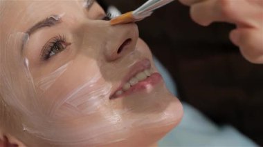 Yetenekli bir kadın kozmetikçi, doğal güzelliğini arttırmak için gülümseyen beyaz bir müşteriye nazikçe yüz maskesi takar. Yakın çekim, hassas fırça darbelerini ve müşterilerin sakin ifadesini yakalar.